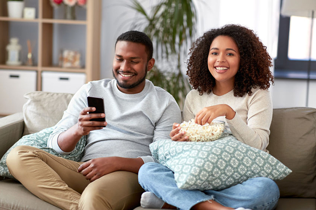 A man and woman sitting on a couch, enjoying popcorn while watching television together.