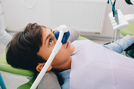 The image shows a young person sitting in a dental chair with their eyes closed, wearing a medical face mask and a device that appears to be a dental impression tray, possibly during a dental procedure.
