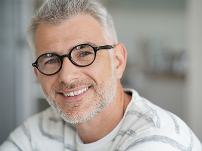 The image features a man with glasses smiling at the camera, appearing to be middle-aged with short hair, wearing a white shirt with a patterned design.