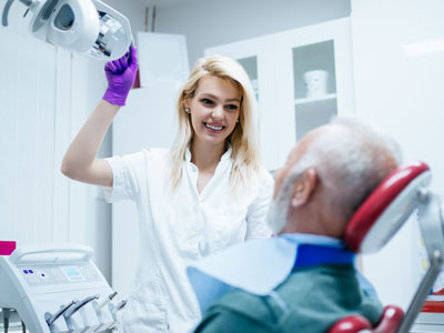 A woman wearing a white lab coat stands next to an older man in a dentist's chair, holding a dental device with a smile on her face.