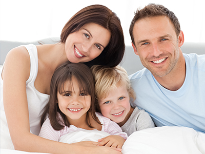 The image shows a family of five people, including two adults and three children, sitting closely together on a bed with smiles on their faces.