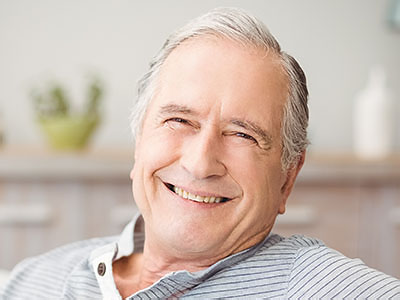 A man with gray hair sitting comfortably in his home, smiling broadly at the camera.