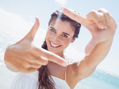 The image features a woman with long hair smiling at the camera while making a peace sign with her fingers. She is outdoors, wearing a white top, and appears to be enjoying a sunny day by the water.