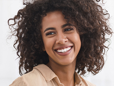 A woman with curly hair and a radiant smile, wearing a beige top, stands against a white background.