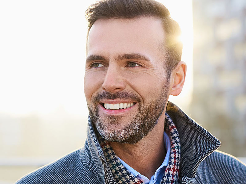 The image features a man with facial hair smiling towards the camera, wearing a checkered jacket and a blue shirt, against a blurred background of a cityscape during daylight.